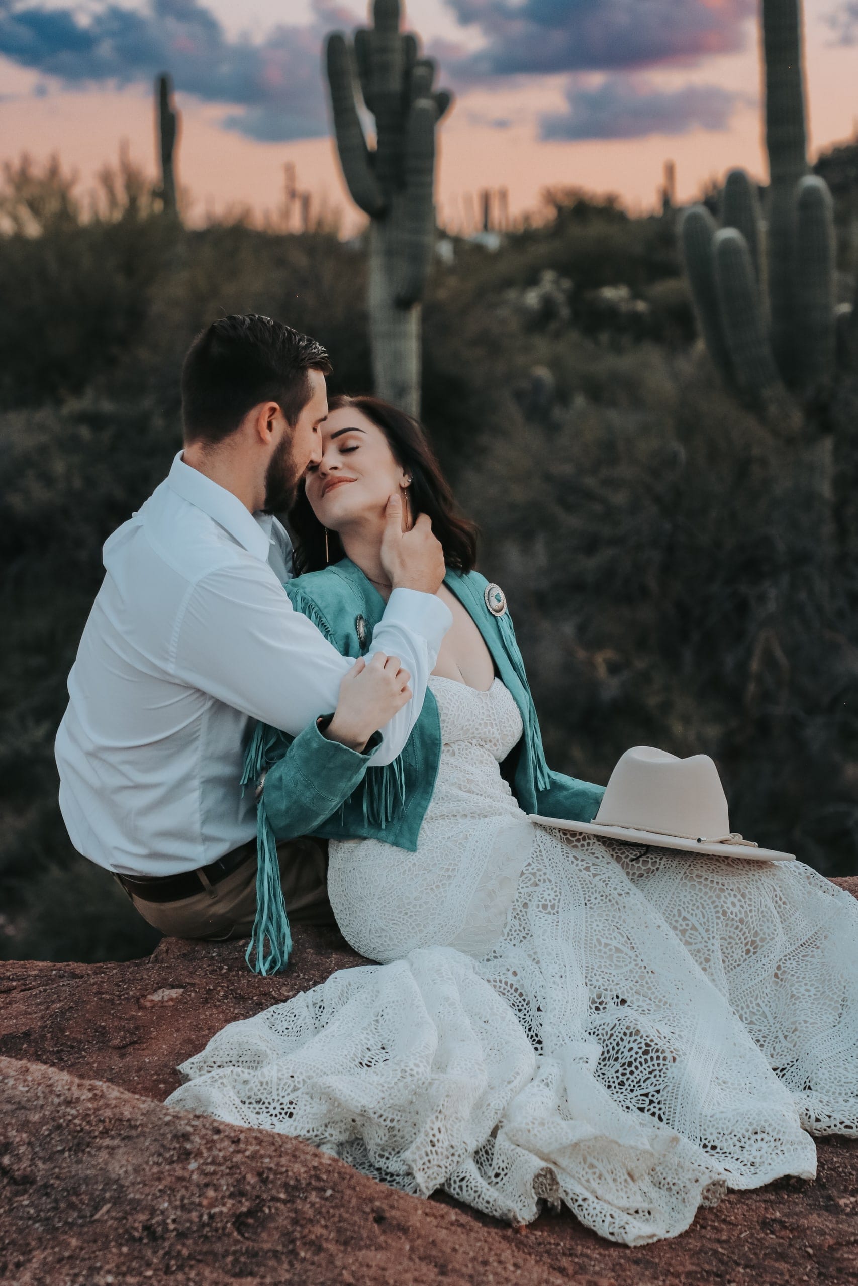 Couple embraces among saguaros at dusk, bride in a turquoise fringe jacket and lace gown