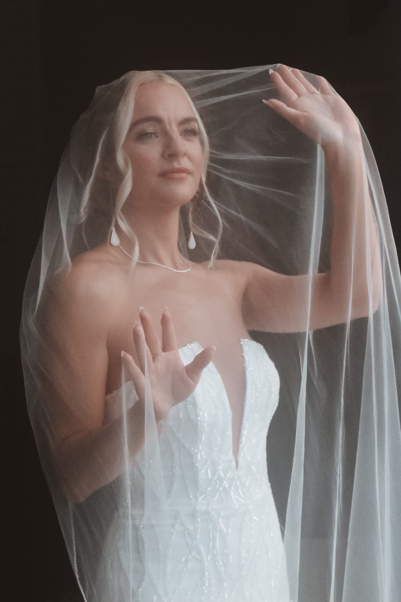Bride lifts her veil against a dark backdrop