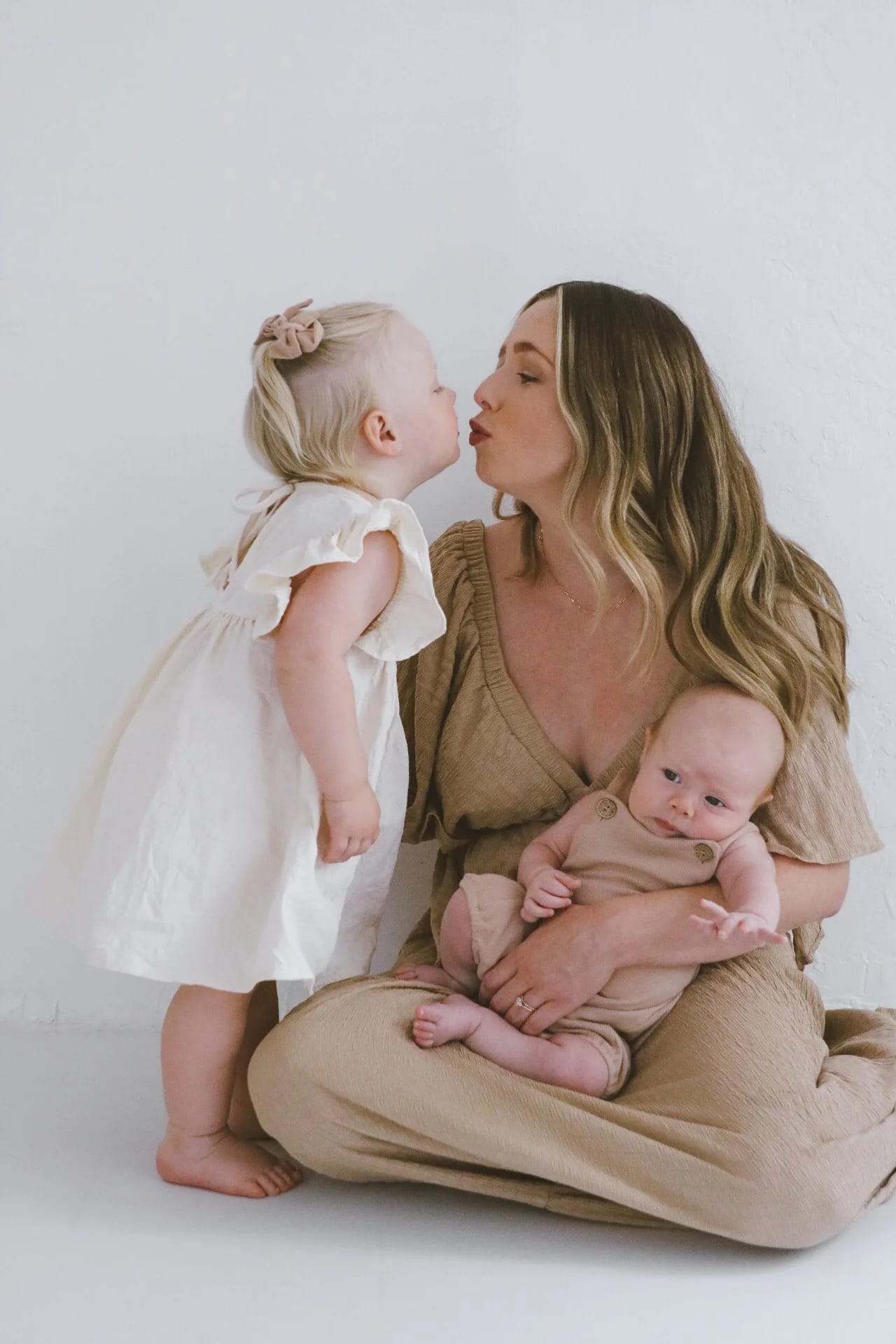 Mother holds her newborn while her toddler kisses her cheek
