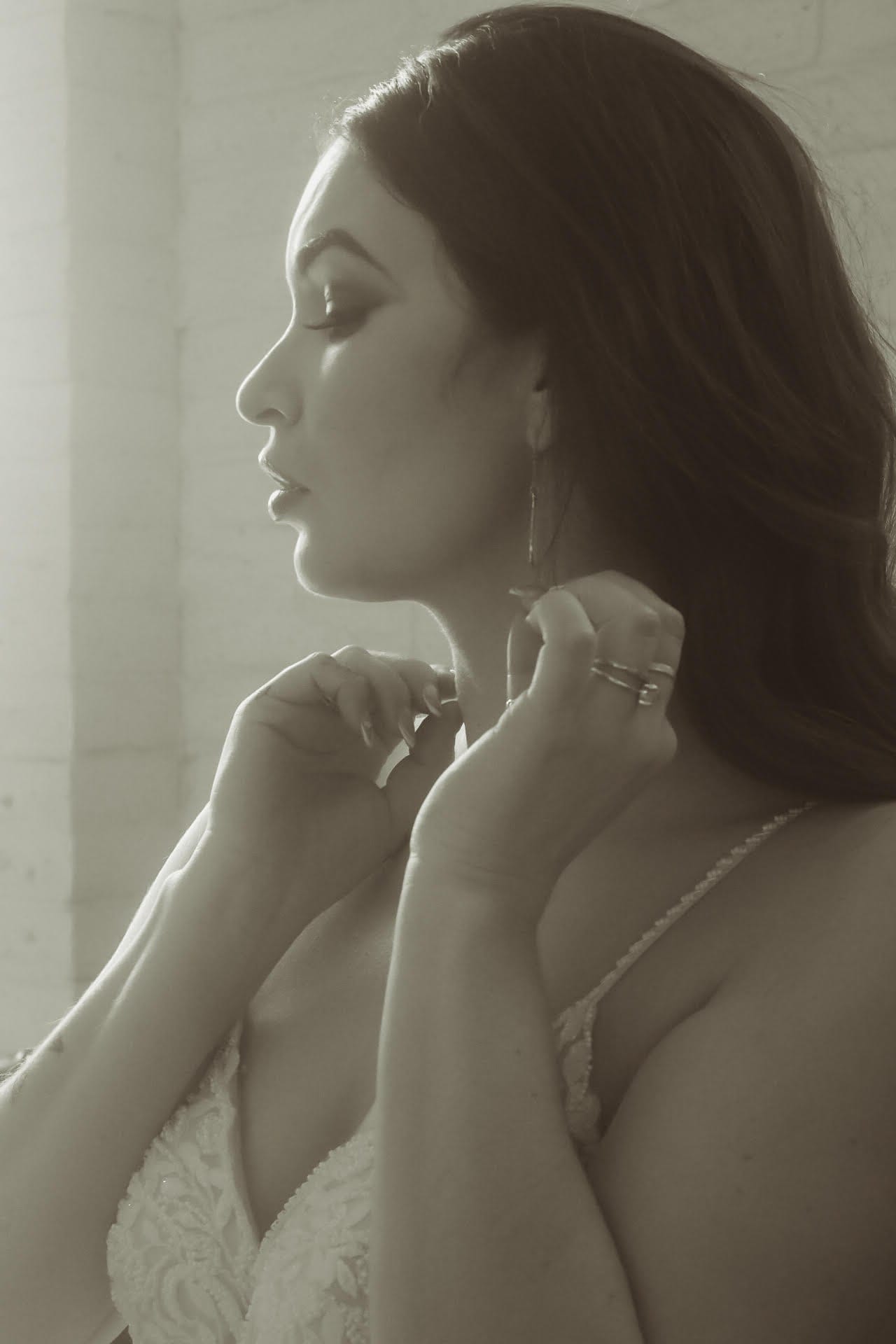 Black and white bridal portrait, bride fastening her earring by the window
